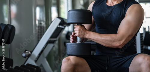 Selective focus of sport man bodybuilder resting with heavy dumbbells after done workout exercise at gym. Muscular man in sportswear sitting after do weightlifting for strength and firm muscular.