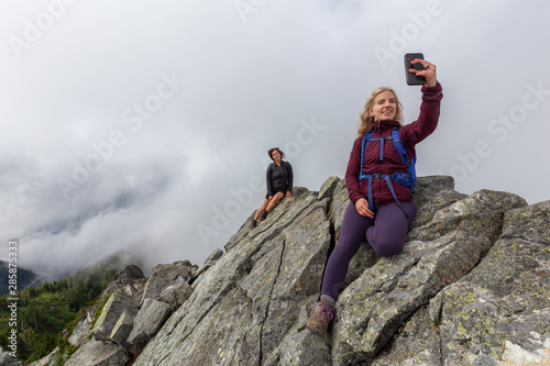 Young Adventurous Girl on top of a rugged rocky mountain taking a selfie during a cloudy summer morning. Taken on Crown Mountain, North Vancouver, BC, Canada.