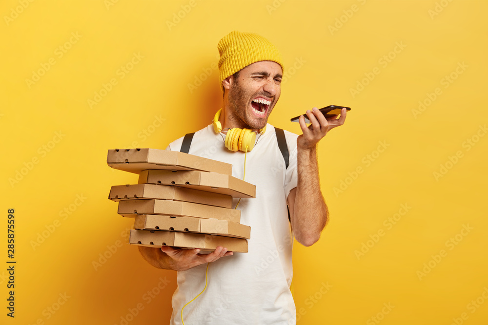 Photo of annoyed pizza man courier screams angrily at smartphone, has annoying conversation with client, holds pile of carton boxes, wears hat and white t shirt, isolated on yellow background Photo of annoyed pizza man courier screams angrily at smartphone, has annoying conversation with client, holds pile of carton boxes, wears hat and white t shirt, isolated on yellow background