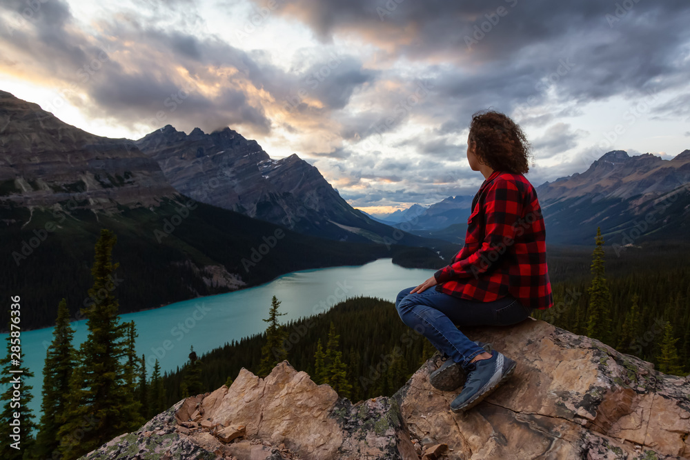 Adventurous girl sitting on the edge of a cliff overlooking the beautiful Canadian Rockies and Peyto Lake during a vibrant summer sunset. Taken in Banff National Park, Alberta, Canada.