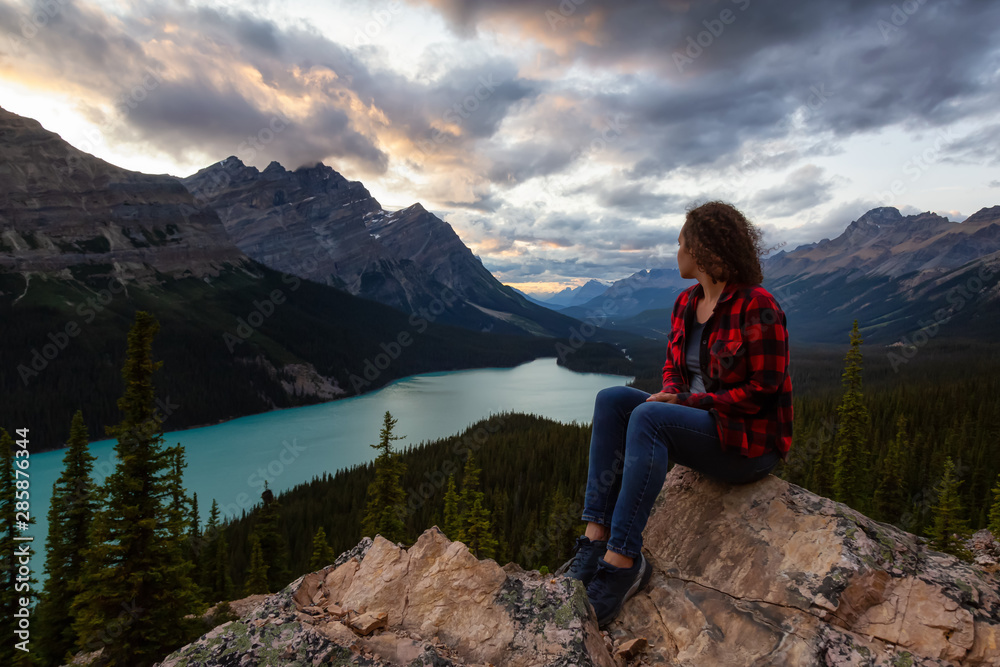 Adventurous girl sitting on the edge of a cliff overlooking the beautiful Canadian Rockies and Peyto Lake during a vibrant summer sunset. Taken in Banff National Park, Alberta, Canada.