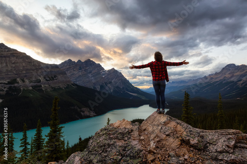 Adventurous girl with open arms standing on the edge of a cliff overlooking the beautiful Canadian Rockies and Peyto Lake during a vibrant summer sunset. Taken in Banff National Park, Alberta, Canada.