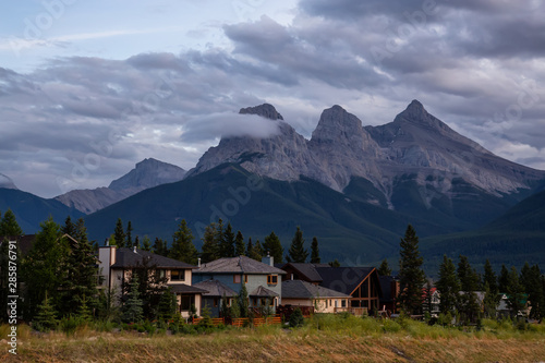 Beautiful view of Residential Homes with Canadian Rocky Mountains in the background during a cloudy summer sunset. Taken in Canmore, Alberta, Canada.