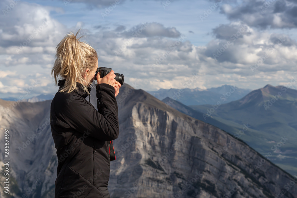 Adventure female Photographer is taking pictures on top of a rocky mountain during a cloudy day. Taken from Mt Lady MacDonald, Canmore, Alberta, Canada.