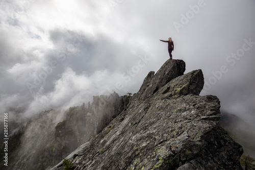 Adventurous Girl pointing finger on top of a rugged rocky mountain during a cloudy summer morning. Taken on Crown Mountain  North Vancouver  BC  Canada.