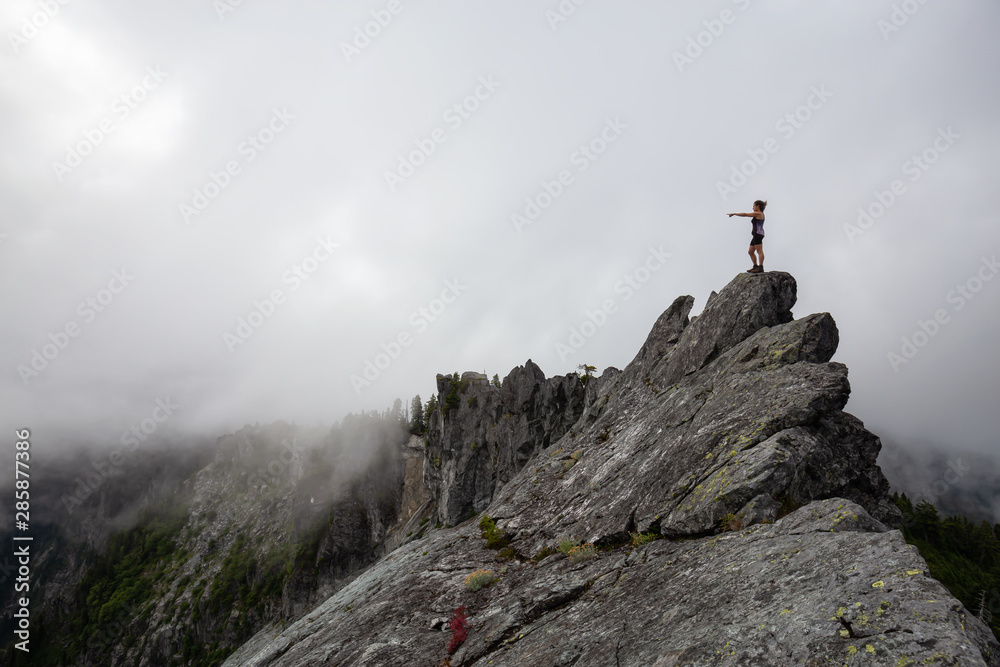 Adventurous Girl pointing finger on top of a rugged rocky mountain during a cloudy summer morning. Taken on Crown Mountain, North Vancouver, BC, Canada.