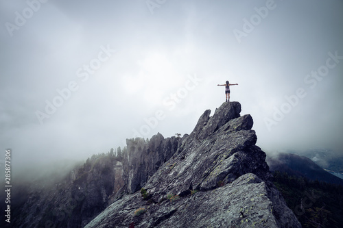 Adventurous Girl with Open Arms on top of a rugged rocky mountain during a cloudy summer morning. Taken on Crown Mountain, North Vancouver, BC, Canada.