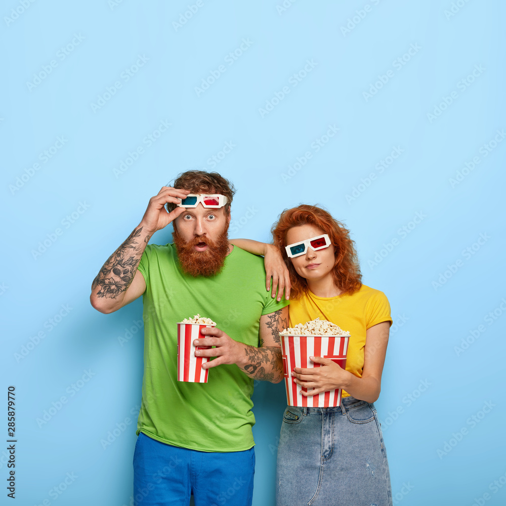 Obraz premium Vertical shot of shocked man stares with surprise, takes off 3d glasses, sad bored woman leans at shoulder, spend free time at cinema, eat tasty popcorn, pose indoor. People and entertainment