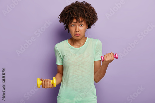 Puzzled African American woman looks surprisingly, holds two dumbbells, works on muscles, wears t shirt, likes fitness and sport, isolated on purple studio wall. Lady with weights trains in gym