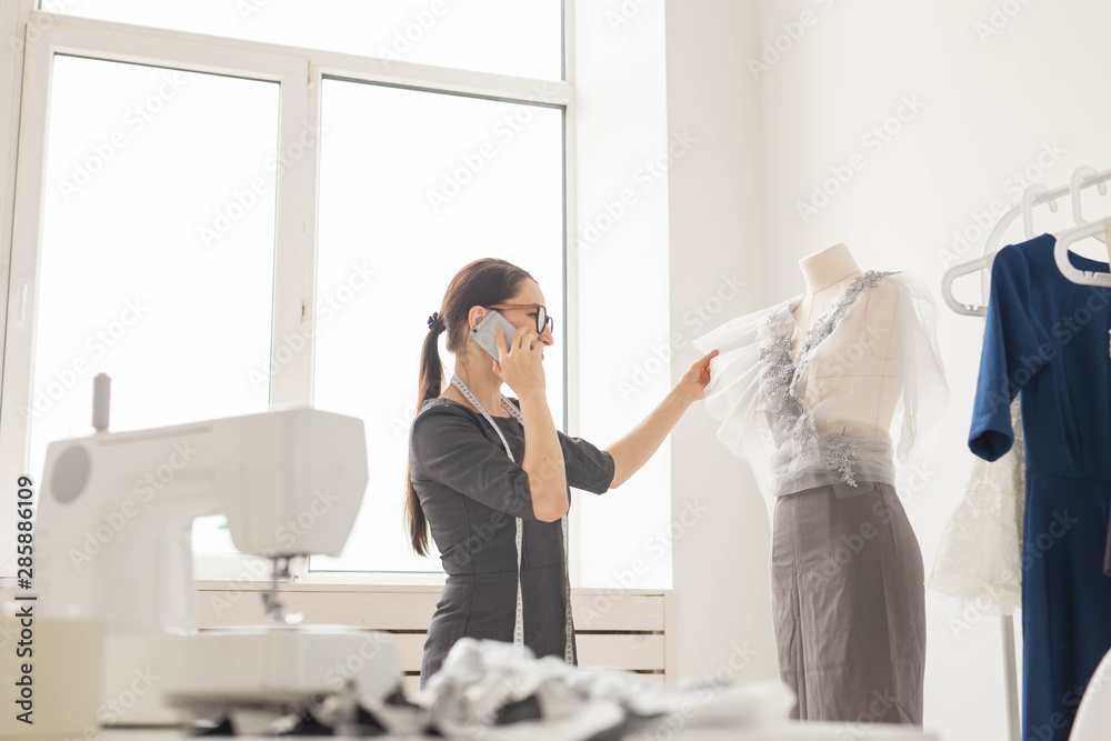 Naklejka premium Dressmaker, tailor, fashion and showroom concept - beautiful young designer is talking on the mobile phone and smiling while examining her work in dressmaking studio