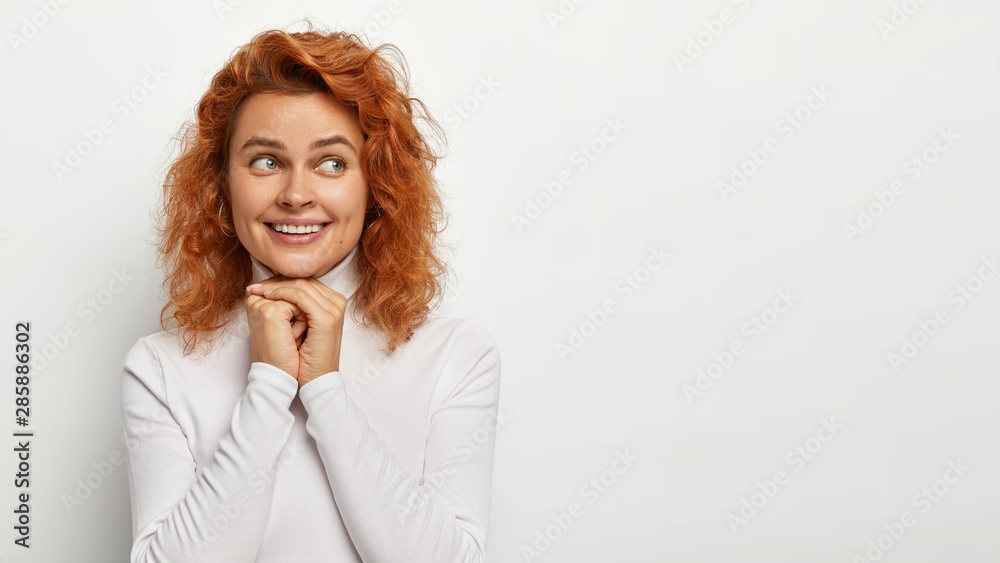 Indoor shot of pleasant looking ginger woman keeps hands under chin, looks positively aside, has gentle smile, wears casual jumper, isolated on white studio wall with blank space. Happiness concept Indoor shot of pleasant looking ginger woman keeps hands under chin, looks positively aside, has gentle smile, wears casual jumper, isolated on white studio wall with blank space. Happiness concept