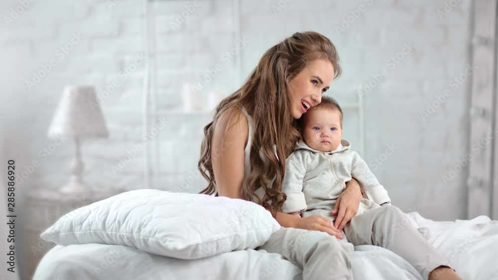 Pretty woman relaxing with cute baby sitting on bed at white bedroom feeling positive emotion Pretty woman relaxing with cute baby sitting on bed at white bedroom feeling positive emotion