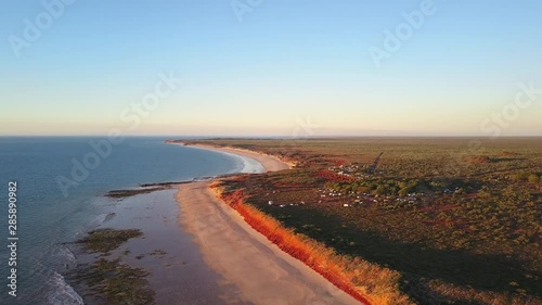 Scenic panoramic aerial drone view of camping area at remote beach in Western Australia, flying along ocean, red rocky cliffs, with blue sky, sunset and horizon on a summer sunny day.