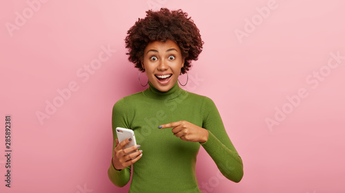 Just look at my electonic device! Cheerful beautiful African American woman points at mobile phone, surprised pleasantly with its innovative functions, gazes happily, models indoor in casual wear photo