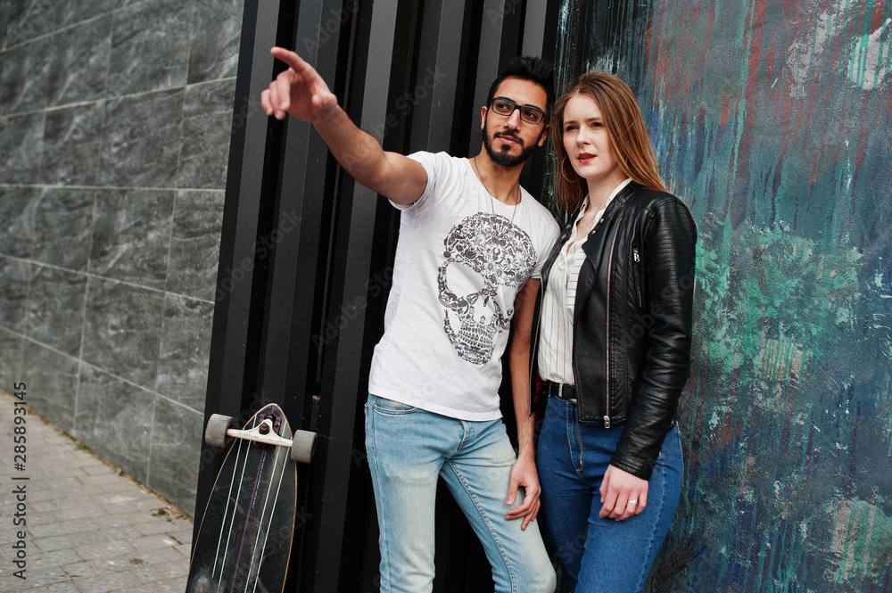 Cool multiracial couple posing against wall with longboard. Arab boyfriend show something to his european girl.