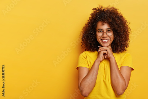 Lovely positive Afro woman has curly hair, healthy skin, keeps hands together under chin, glad to hear pleasant comment about her work, wears yellow t shirt, models indoor. Human emotions concept