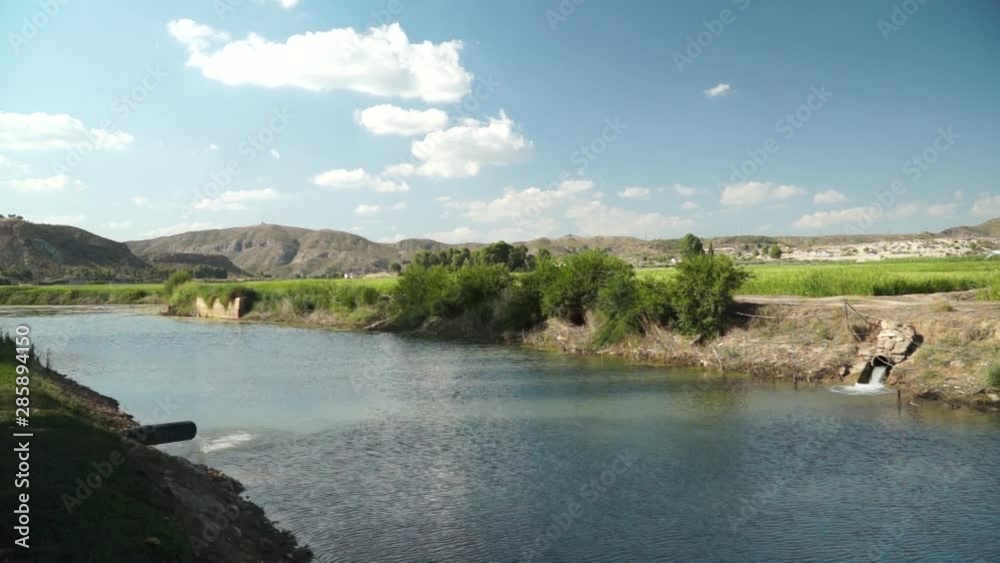 Static shot of a flowing river with the mountains in the background under the blue skies and clouds Static shot of a flowing river with the mountains in the background under the blue skies and clouds