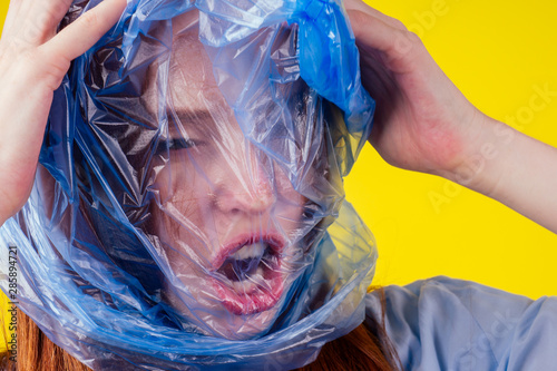 Fotografie redhaired ginger european woman suffocation head in blue polyethylene package in studio yellow background
