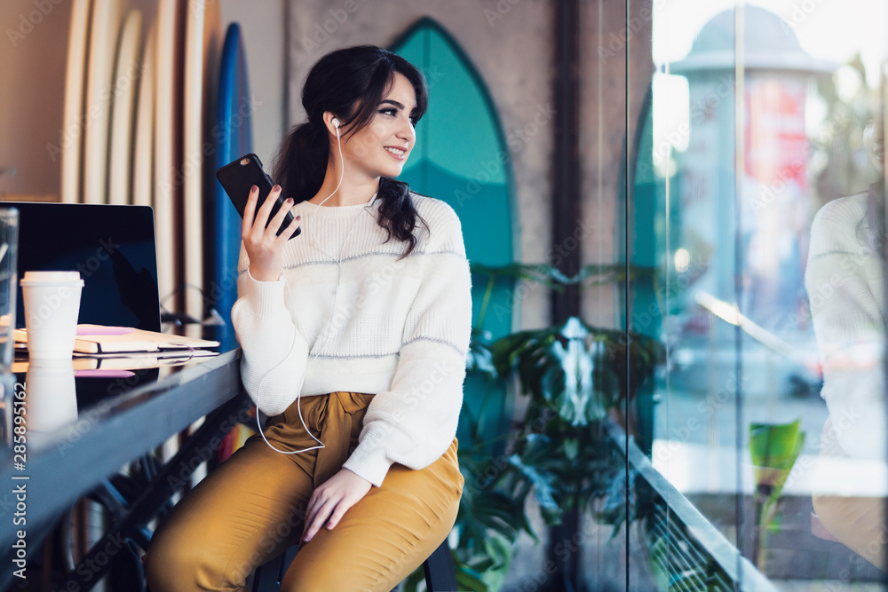 Young smiling woman sitting in cafe at table,looking out window, holding smartphone, listening to music on headphones. Lifestyle. Young smiling woman sitting in cafe at table,looking out window, holding smartphone, listening to music on headphones. Lifestyle.