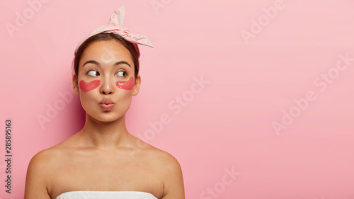 Quadro su tela Beautiful young korean female model wears eye patches for fresher look, cares about skin, looks aside, keeps lips folded, isolated over pink background with blank space for promo