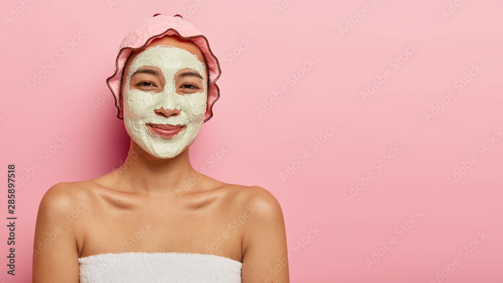 Good looking gentle Asian girl has clay facial mask, smiles happily, wears bathcap on head, wants to have clean fresh skin, visits cosmetologist in spa salon, isolated on rosy studio wall, free space Good looking gentle Asian girl has clay facial mask, smiles happily, wears bathcap on head, wants to have clean fresh skin, visits cosmetologist in spa salon, isolated on rosy studio wall, free space