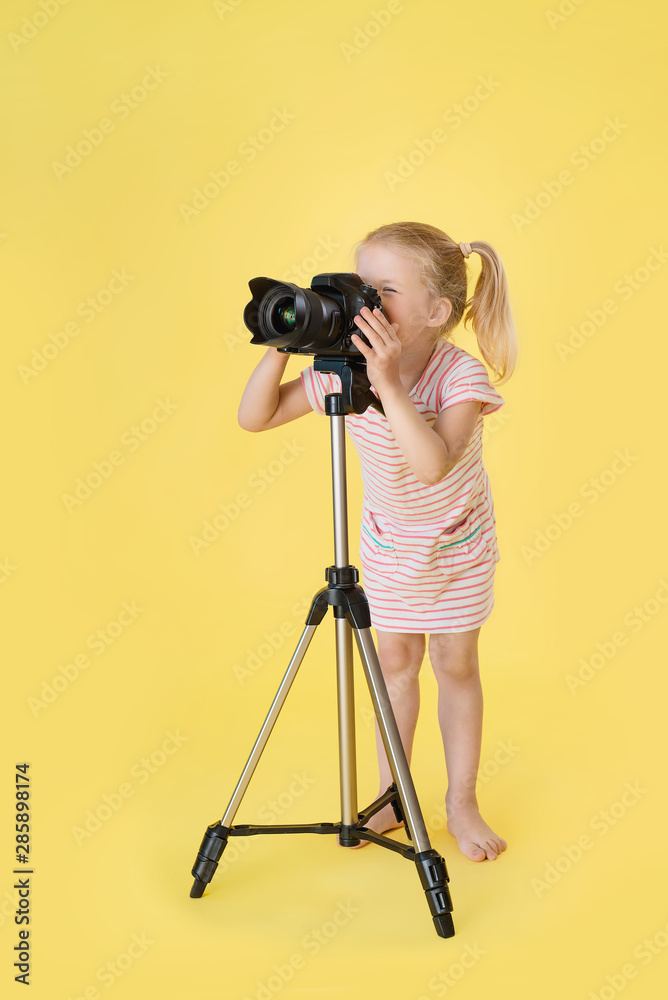 Little pretty girl on yellow background shooting with professional photo camera and tripod
