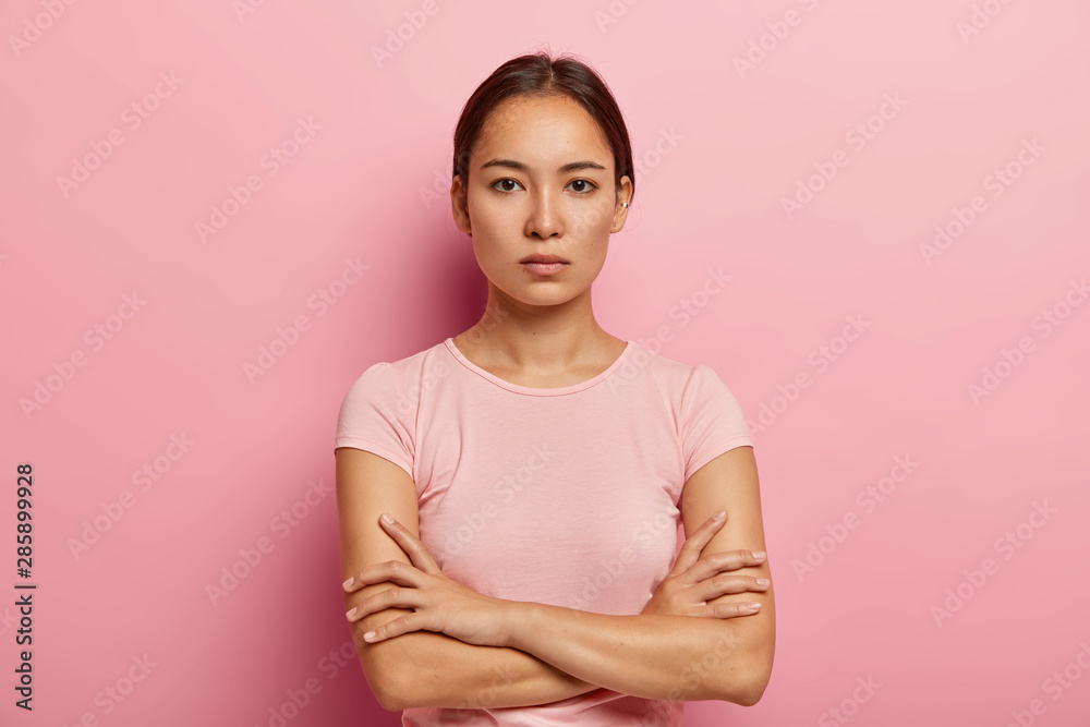 Headshot of serious korean woman looks with calm face expression, keeps arms folded, has healthy fresh skin, wears rosy t shirt, stands indoor. Beautiful Asian girl has confident gaze at camera Headshot of serious korean woman looks with calm face expression, keeps arms folded, has healthy fresh skin, wears rosy t shirt, stands indoor. Beautiful Asian girl has confident gaze at camera