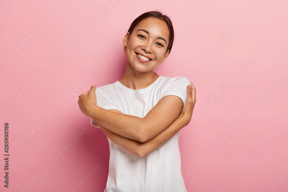Love yourself. Pretty glad Asian girl embraces herself, feels comfort and care, tilts head, wears white t shirt, has no make up, isolated over rosy wall, thinks about lover, wants be in his warm arms Love yourself. Pretty glad Asian girl embraces herself, feels comfort and care, tilts head, wears white t shirt, has no make up, isolated over rosy wall, thinks about lover, wants be in his warm arms