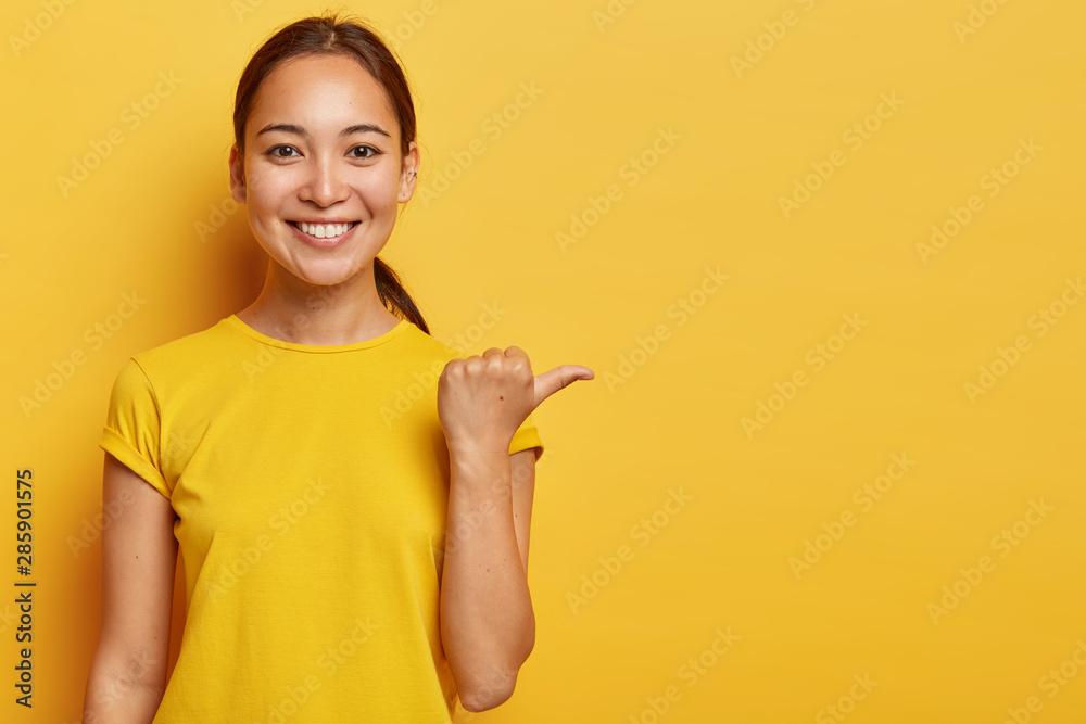 Studio portrait of young cheerful Asian female points away with thumb, happy face expression, demonstrates copy space for advertisement, has pleasant appearance, wears bright yellow clothes. - obrazy, fototapety, plakaty 
