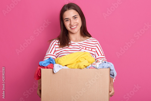 Studio shot of adorable female stands smiling with box full of reusable clothing for poor people isolated over pink background, attractive woman making charity, young lady wearing striped shirt. photo