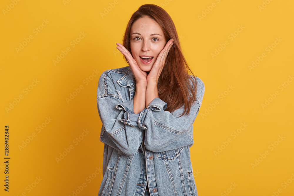 Close up portrait of young girl has surprised facial expression with hands on her cheek. Beautiful young woman holding her mouth opened, dressed denim jacket posing isolated over yellow background. Close up portrait of young girl has surprised facial expression with hands on her cheek. Beautiful young woman holding her mouth opened, dressed denim jacket posing isolated over yellow background.