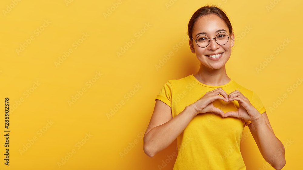 Lovely korean woman with happy smile, shapes heart with both hands, expresses love to you, wears round spectacles and yellow t shirt, says be my valentine flirts with boyfriend poses indoor copy space - obrazy, fototapety, plakaty 