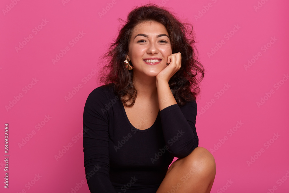 Close up portrait of young cheerful beautiful girl with dark wavy hair dressed black shirt, sitting smiling with teeth against pink wall, looking in camera with happy and relaxed facial expression. Close up portrait of young cheerful beautiful girl with dark wavy hair dressed black shirt, sitting smiling with teeth against pink wall, looking in camera with happy and relaxed facial expression.