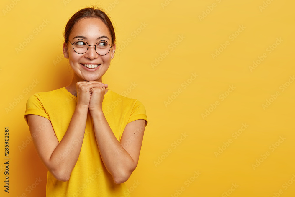 Happy eastern girl looks joyfully aside, holds hands under chin, being delighted and dreamy, focused aside, has dark hair, wears spectacles and t shirt, isolated on yellow studio wall with empty space Happy eastern girl looks joyfully aside, holds hands under chin, being delighted and dreamy, focused aside, has dark hair, wears spectacles and t shirt, isolated on yellow studio wall with empty space