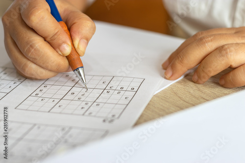 Solve sudoku puzzle with pencil as hobby by senior woman on wooden office desk. Player insert numbers into grid consisting of nine squares subdivided into further nine smaller squares. photo
