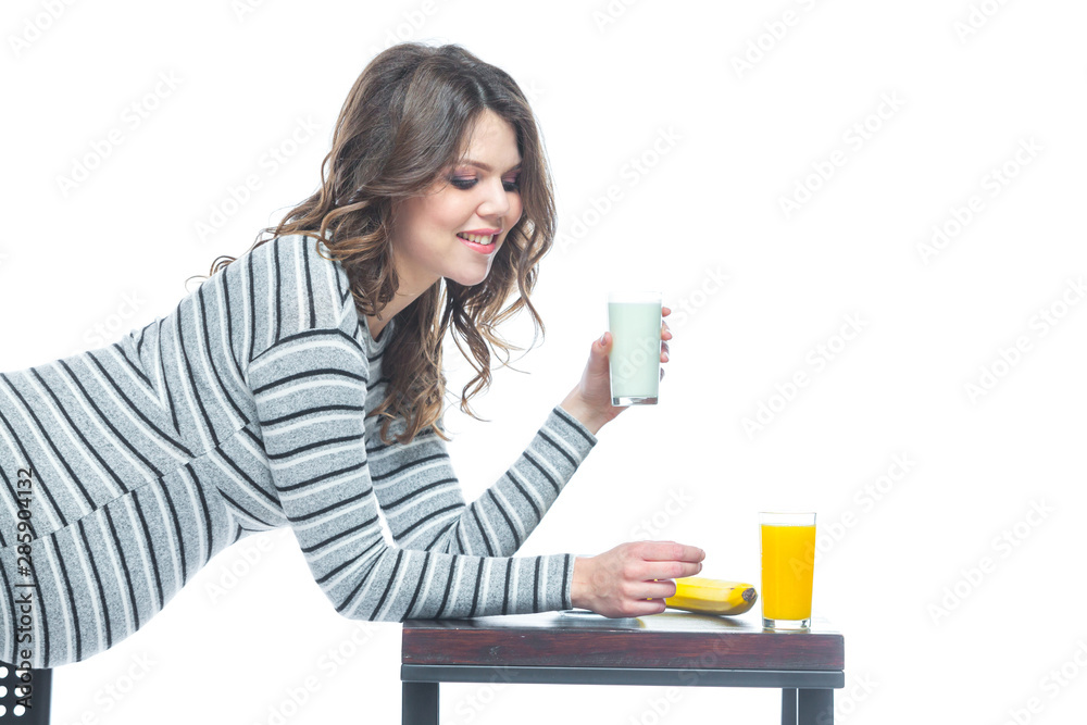 A young pregnant woman sits at a table on which lies a banana, there is a plate with cereal, a glass with kefir and juice. The concept of nutrition during pregnancy. Isolated on a white background. A young pregnant woman sits at a table on which lies a banana, there is a plate with cereal, a glass with kefir and juice. The concept of nutrition during pregnancy. Isolated on a white background.