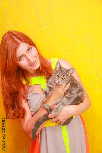 pretty young redheaded girl in long colorful summer dress with her lovely friend grey cat on yellow bright studio background
