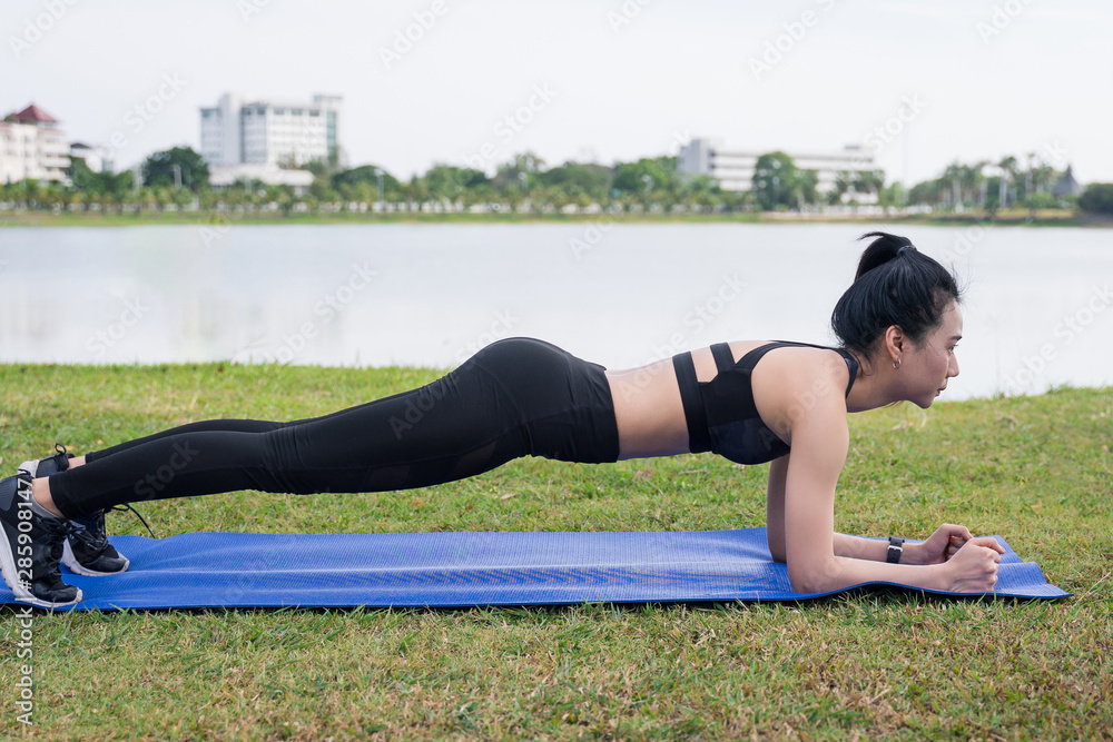 Fototapeta premium Slim beautiful women practices yoga poses on the mat beside a lake at the outdoor park with green grass background. Yoga and life health concept with copy and paste space.