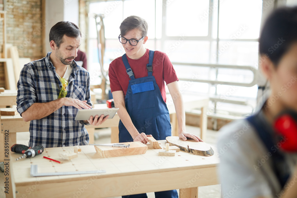 Waist up portrait of mature carpenter using digital tablet while talking to smiling young apprentice in workshop, copy space Waist up portrait of mature carpenter using digital tablet while talking to smiling young apprentice in workshop, copy space