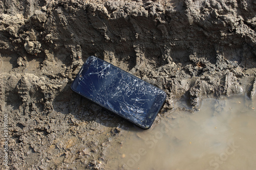 The smartphone with a broken display lies in a dirt, cracks and damage to the device.
