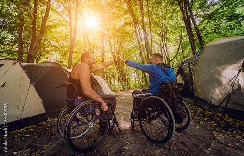 Two disabled men resting in a campsite with friends. Wheelchair in the forest on the background of tents