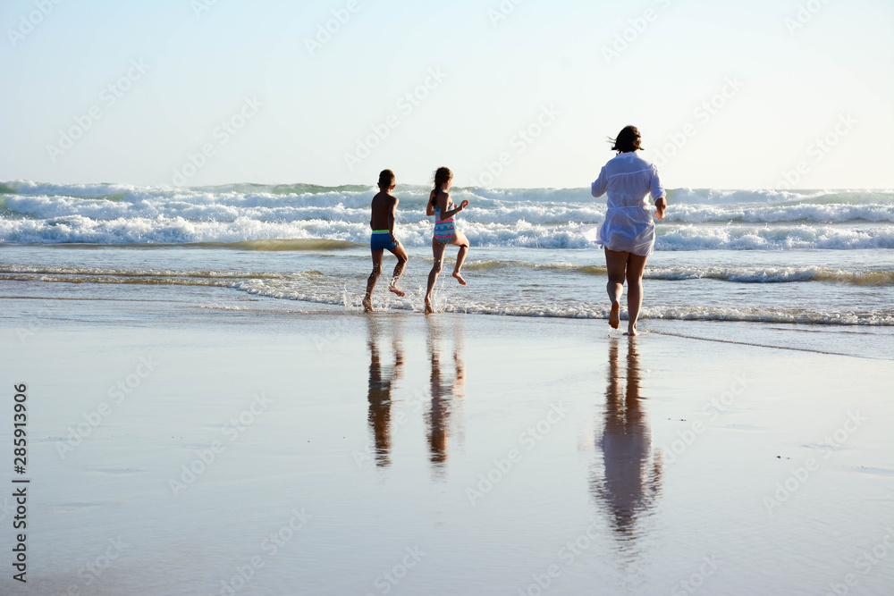 Mother with son and daughter running into the ocean before sunset. Reflection on sand with water.