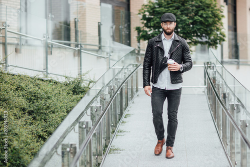 A bearded, serious, stylish man walking on the streets of the city near modern office centre and drinks coffee. Copy space.