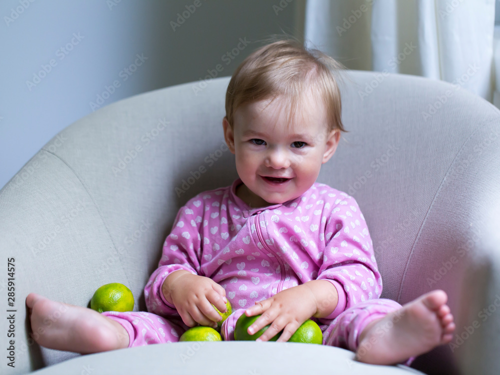 Full length horizontal shot of adorable fair smiling toddler girl wearing pink pyjamas sitting in armchair playing with limes Full length horizontal shot of adorable fair smiling toddler girl wearing pink pyjamas sitting in armchair playing with limes