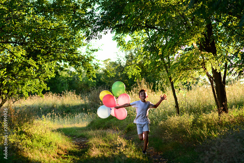 The boy runs through the forest with colorful balloons before sunset. A school boy is chasing through a forest with colorful balloons in a white t-shirt with rays of sun.