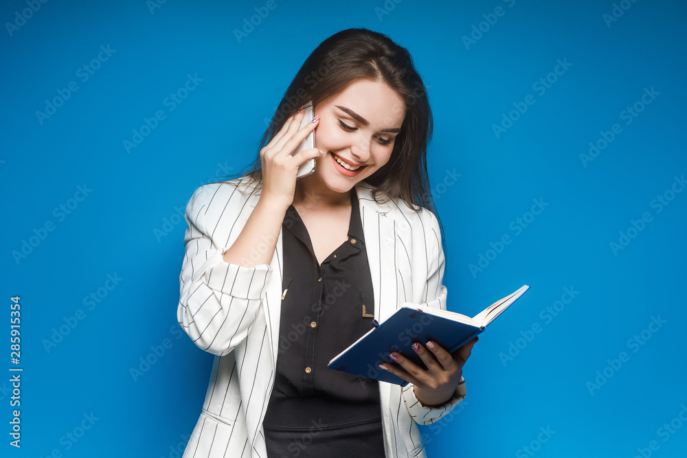 Brunette, attractive businesswoman speak by phone with copybook isolated on blue background. Brunette, attractive businesswoman speak by phone with copybook isolated on blue background.