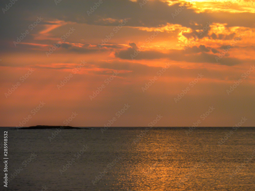 A beautiful sunset over the sea, The sun beams are reflecting itself in calm and wave-less surface of the sea. Sky is exploding with yellow and orange. There is an island in the back.