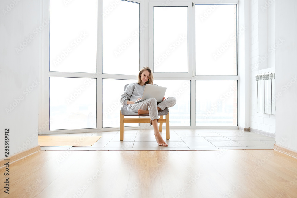 Fototapeta premium Woman in armchair working on laptop near panoramic window