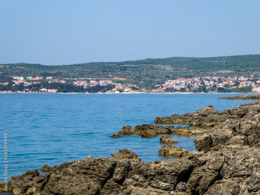 A view on a shallow water in a bay. Sea water is calm and shining with many shades of blue. The rocks striking out of the sea water. In the back there is a big city located by the shore. Clear day