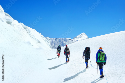 Group of equipped climbers tied with a rope ascent by snowy slope crossing snowfield with crevasses on a way to summit in snowy mountains.